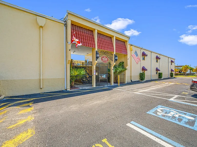 Inviting apartment entrance with vibrant red accents, American and state flags, lush greenery, and ample parking.