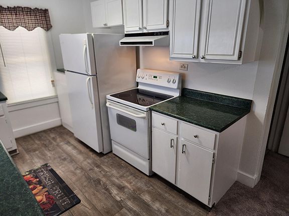 Kitchen showing stove with vent hood light and refrigerator. Microwave (if needed) on other side of refrigerator. Gray flooring.