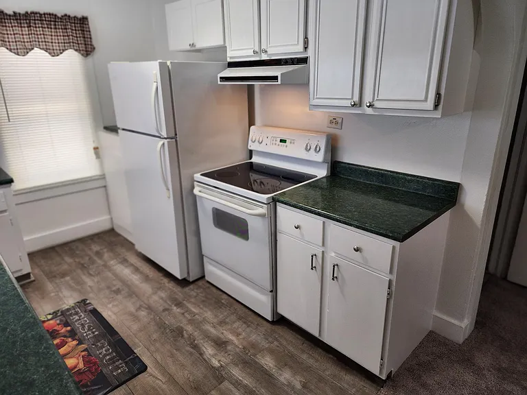 Kitchen showing stove with vent hood light and refrigerator. Microwave (if needed) on other side of refrigerator. Gray flooring.