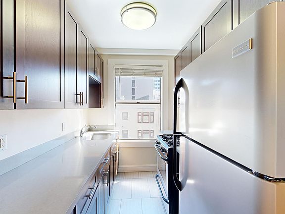 A narrow kitchen with wood cabinets and a stainless steel fridge. Bright light comes through a window above a modern stove, creating an inviting atmosphere.