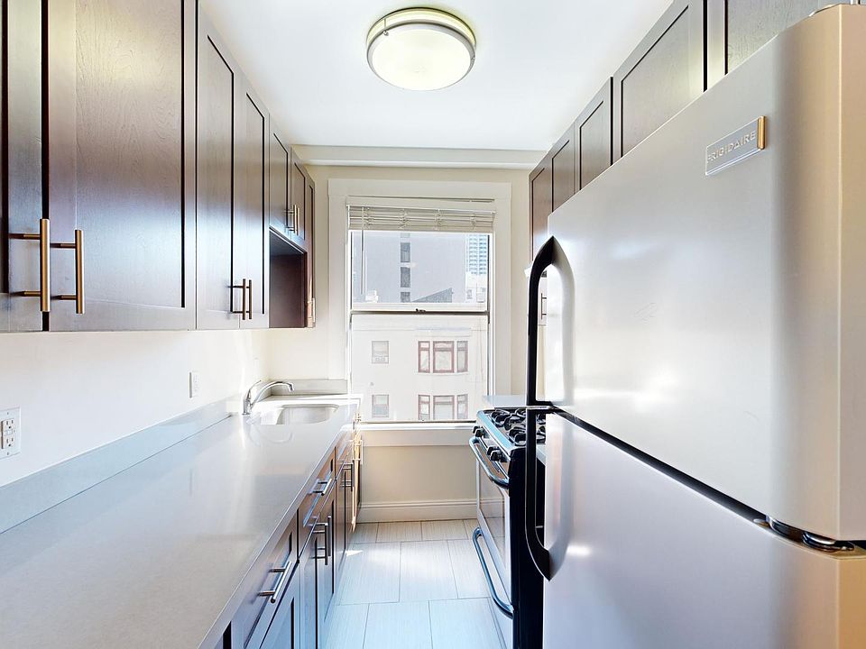A narrow kitchen with wood cabinets and a stainless steel fridge. Bright light comes through a window above a modern stove, creating an inviting atmosphere.