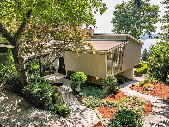 A broader view of the home's entry showcases the unique elevated structure and angular roof lines. The floating staircase, large picture windows, and tiered landscaping emphasize the home's integration with its wooded surroundings.