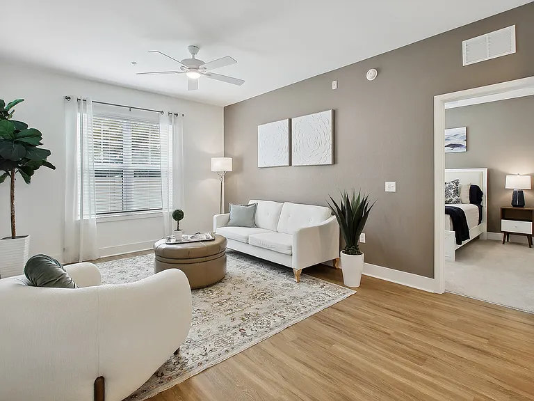 Bright and inviting living room with modern decor, featuring a cozy white sofa, sleek armchair, and a stylish ottoman table on a textured rug. Abundant natural light fills the room through large windows, complemented by elegant furnishings and serene wall