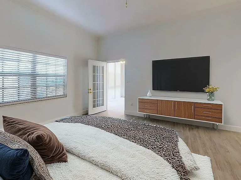 Bright, modern bedroom with sleek wooden flooring, large window with blinds, TV mounted on the wall, and a cozy bed with plush pillows.