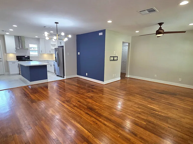 This open-concept living area features beautiful hardwood floors and a modern bright kitchen and a stylish light fixture. A blue accent wall adds a pop of color; recessed dimmer lights and a ceiling fan provides comfort.