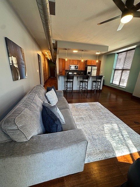 Stylish open-concept living area with a gray sofa, cozy rug, and sleek black chair. The kitchen features warm wood cabinets, stainless steel appliances, and a countertop with barstools. Dark wood floors and exposed ductwork add an industrial-modern vibe.