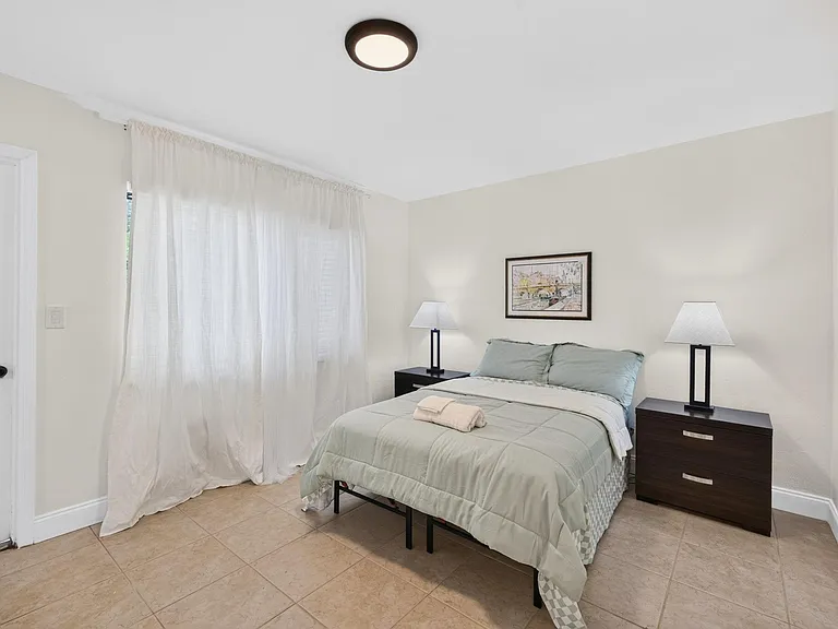 Private bedroom featuring a full bed, matching nightstands with lamps, and a large window with curtains for natural light.
