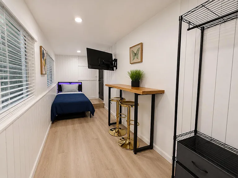 Private bedroom with a twin bed, wall-mounted TV, bar-height desk with stools, and open shelving for storage.