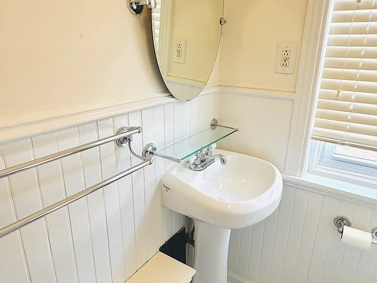 Pedestal sink with chrome fixtures, frameless oval mirror, glass shelf, and towel bars against beadboard paneling.