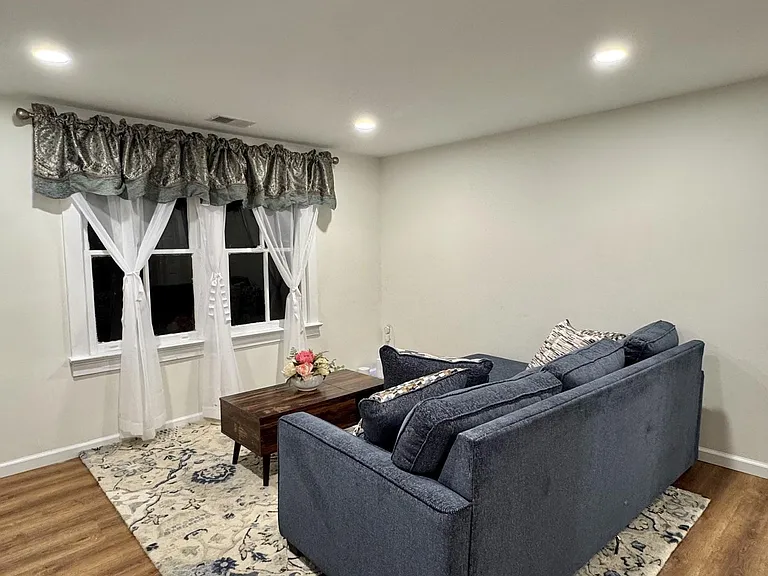 Living room with recessed lighting, a large window with curtains, and seating space anchored by an area rug.