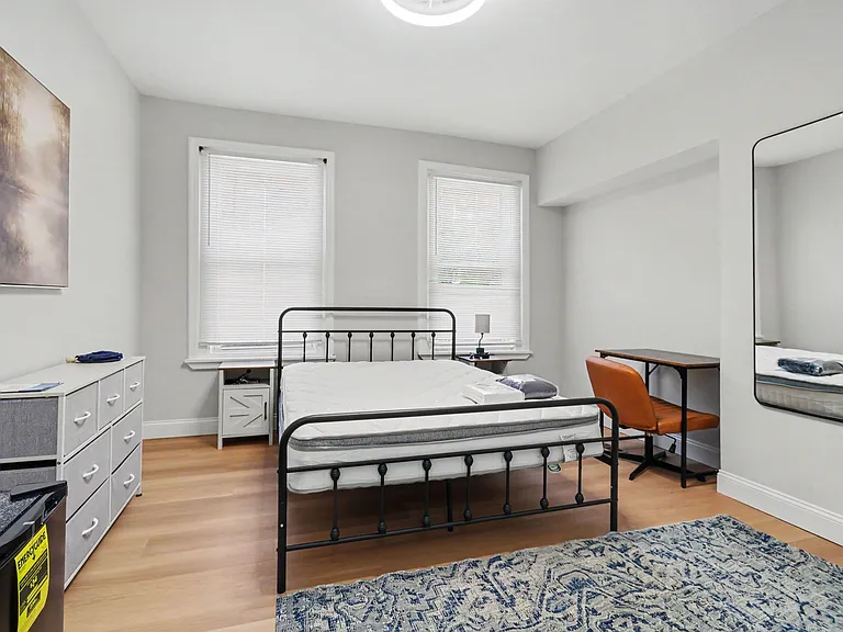 Spacious bedroom with two windows, bed frame, dresser storage, and a dedicated desk setup with full-length mirror.