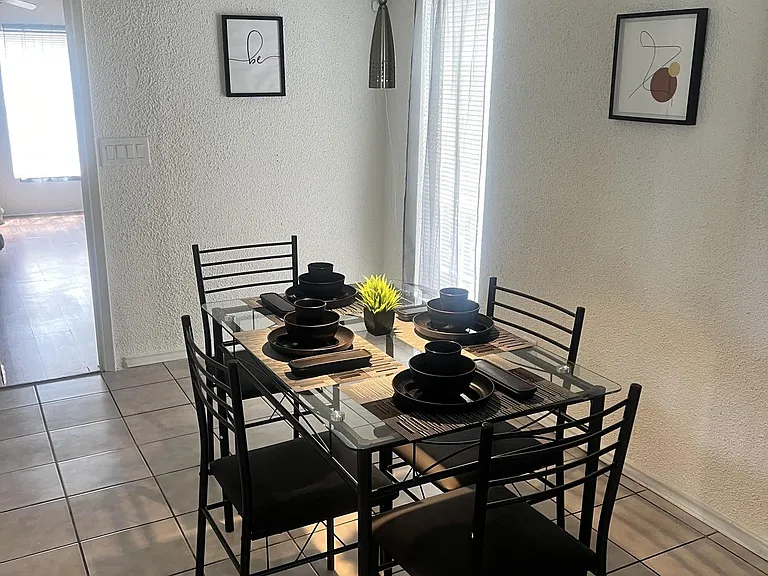 Dedicated dining area with a glass-top table for four, pendant lighting, and a tall window with blinds for natural light.