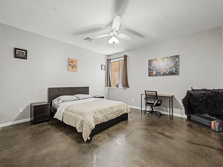 Spacious furnished bedroom with a full bed, nightstands, and a dedicated desk area plus ceiling fan and window curtains.