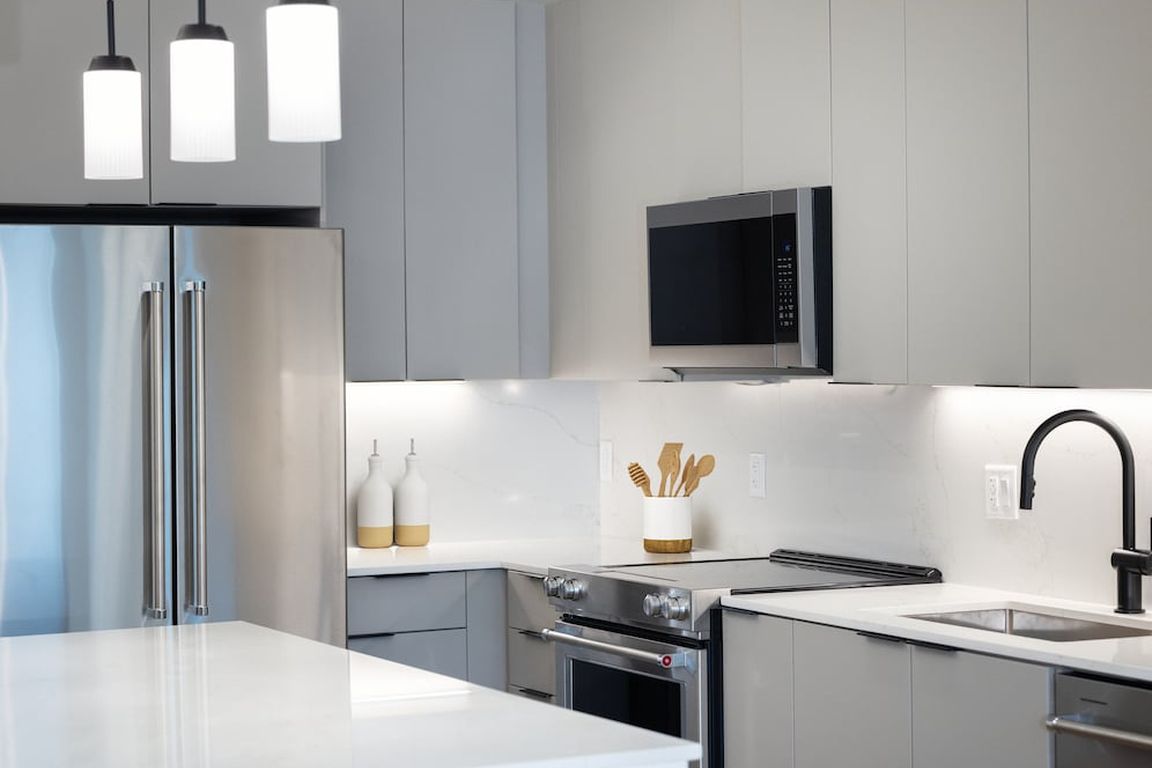 Signature Collection kitchen with stainless steel appliances, light grey cabinetry, marbled quartz countertops and backsplash, under-cabinet lighting, and pendant lighting