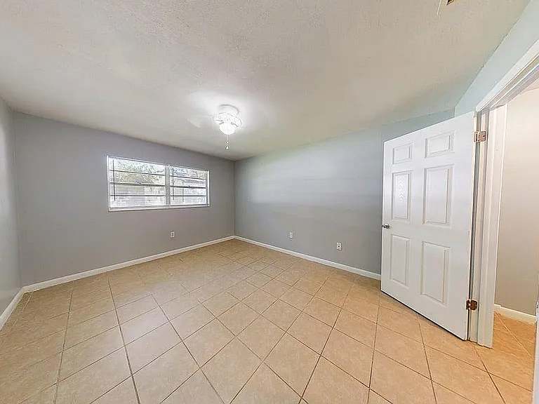 Bright and spacious bedroom with large window, light gray walls, and elegant tile flooring.