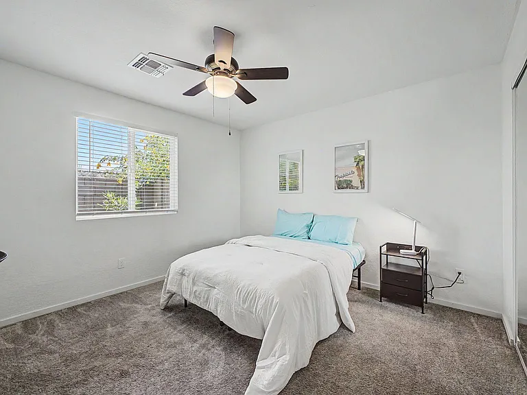 Carpeted bedroom with a full-size bed, bedside table, and ceiling fan, plus a window with blinds and sliding closet doors.