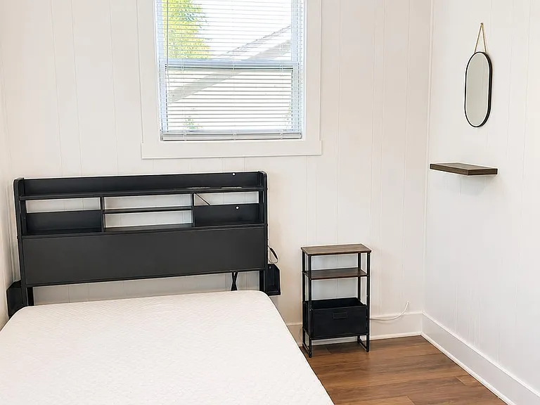 Clean bedroom with a large window, ceiling fan, and bed with headboard shelving plus a nightstand for easy storage.