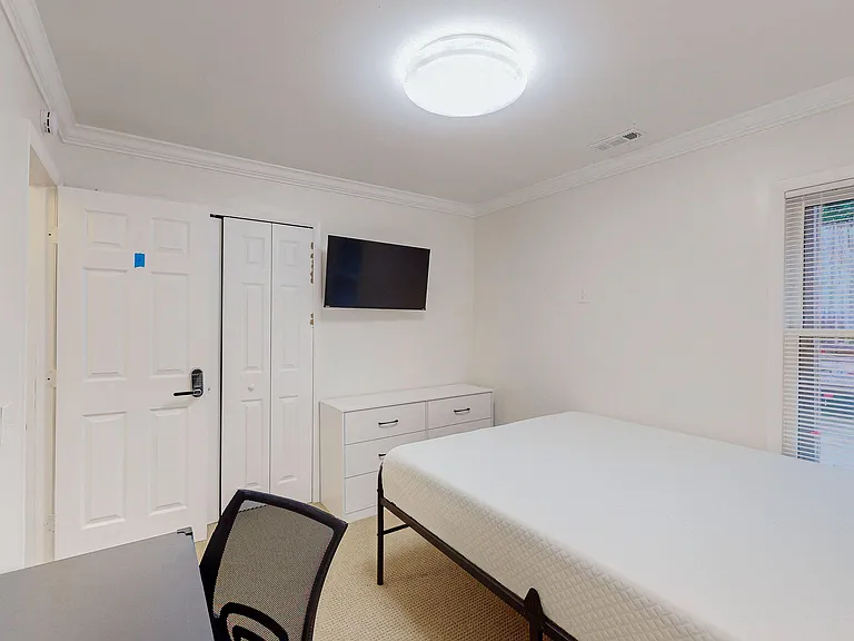 Furnished bedroom with a bed, dresser storage, wall-mounted TV, and a window with blinds for privacy and natural light.