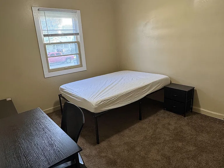 Furnished bedroom with a full bed, nightstand, and desk setup, plus a window with blinds for natural light.