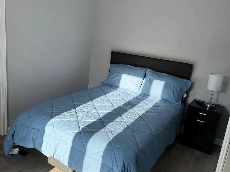 Furnished bedroom with a full-size bed, bedside table and lamp, plus durable flooring and natural light from the window.