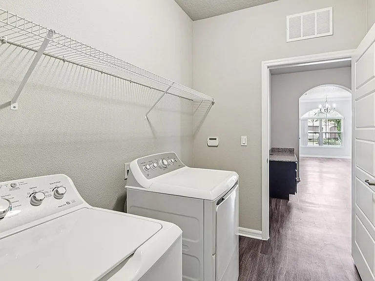 Modern laundry room with sleek white washer and dryer, featuring open shelving and view into sunlit living area with large window.