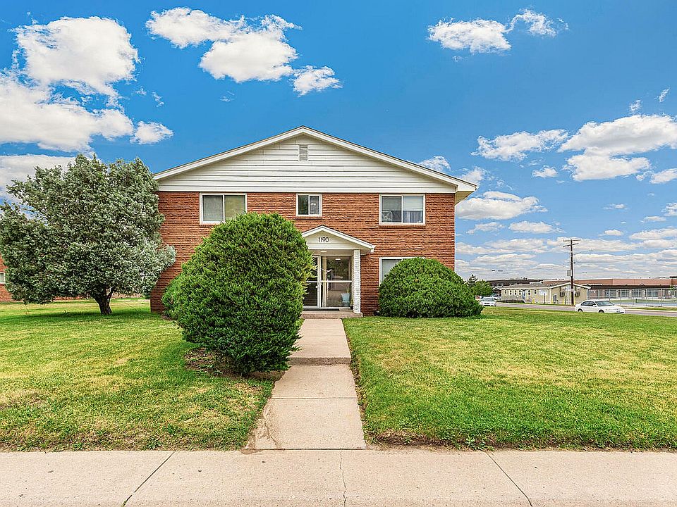 Charming red brick apartment building surrounded by lush greenery and a clear blue sky, offering a welcoming entrance and serene neighborhood views.