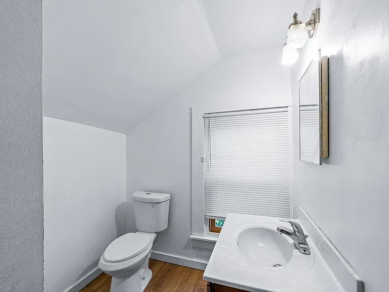 Compact bathroom featuring a single-sink vanity, toilet, overhead lighting, and a window with blinds for ventilation.