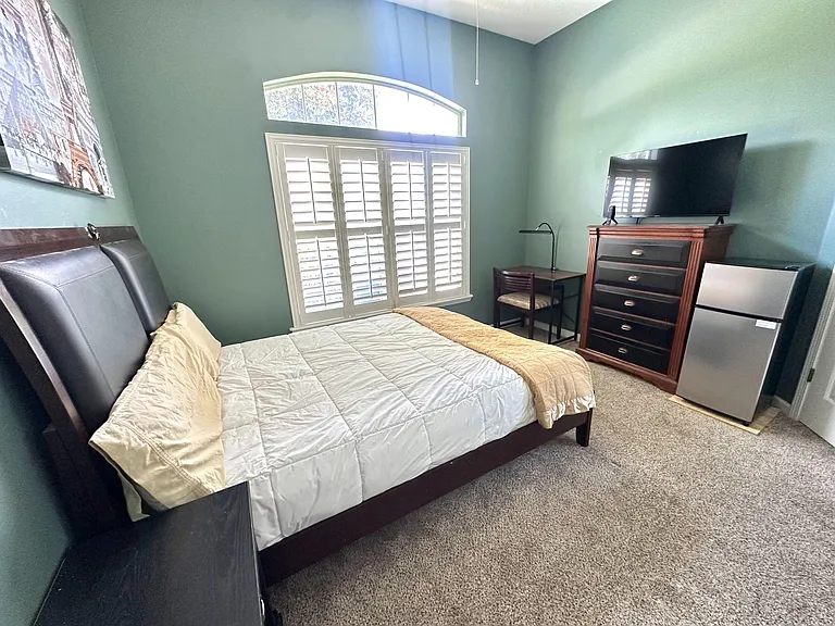Furnished bedroom with a comfortable queen bed, dedicated desk, dresser, and mini fridge, plus large windows with shutters.