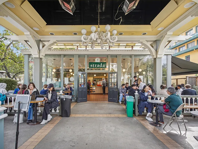 Bustling outdoor cafe with a large glass entrance framed by columns, filled with people seated at tables