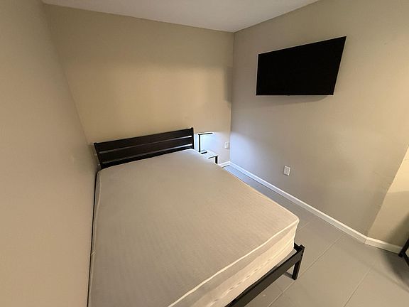 Furnished bedroom with a full bed, nightstand lighting, and a wall-mounted TV for convenient relaxing and streaming.