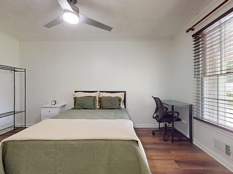 Furnished bedroom with a full bed, large window with blinds, work desk and chair, and open shelving for added storage.