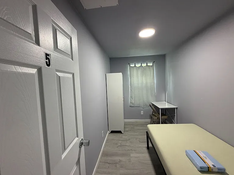 Compact furnished bedroom with bed, wardrobe, and a small desk area, plus a window with curtains and durable flooring.