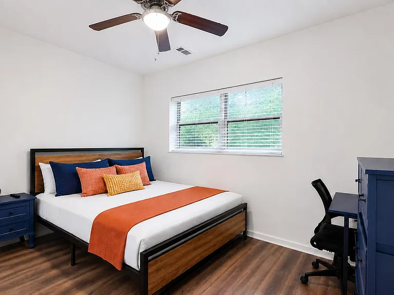 Furnished bedroom with a full bed, nightstand, and dedicated desk area, plus a wide window and ceiling fan for comfort.