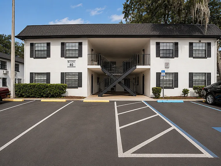 Charming two-story apartment building with classic shutters, surrounded by lush greenery and ample parking space.