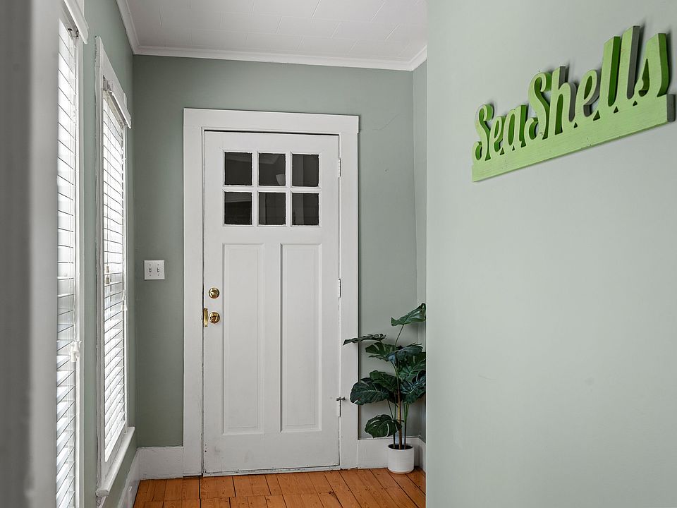 Welcome home! This charming entry area features a classic white door with window panes and original hardwood floors. The walls are a soothing sage green, complemented by nautical/beach-themed decor ("Sea Shells" sign) and bright natural light from the sid