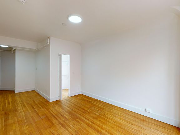 Bright, empty room with hardwood floors. A single doorway leads to another space. Large windows on the right let in natural light.