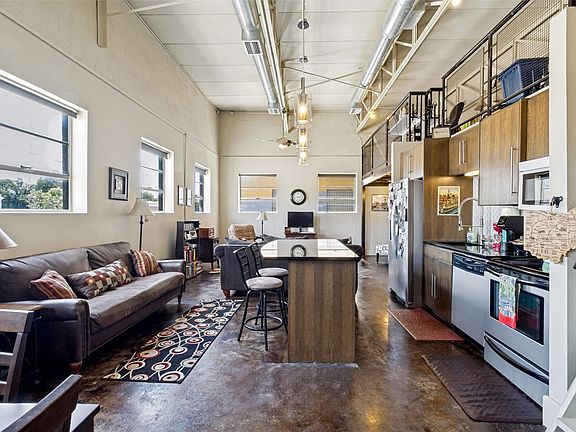 Open-concept living space featuring high ceilings with exposed ductwork, polished concrete flooring, a kitchen island with a dark countertop, stainless steel appliances, and wood-finish cabinetry
