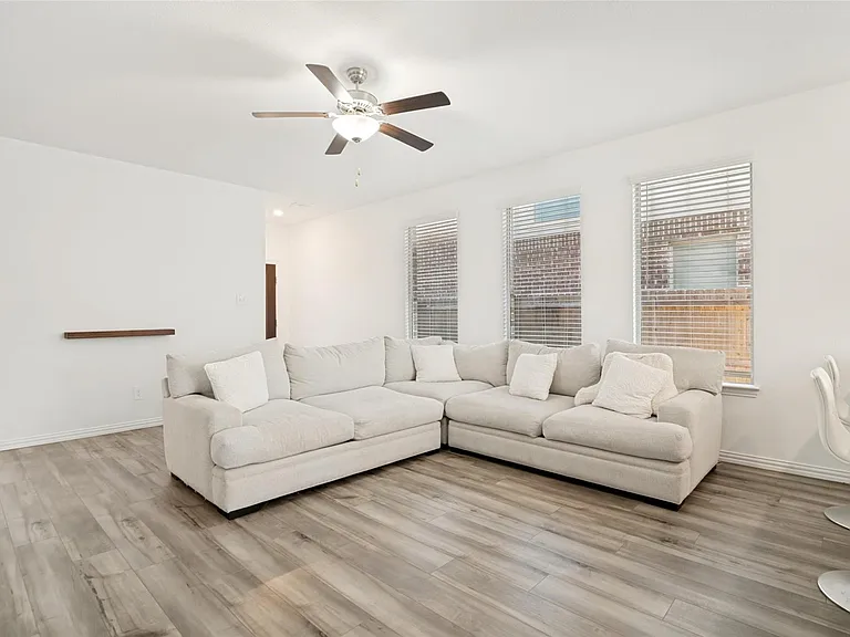 Spacious living area featuring light wood-finish flooring, white walls, and a ceiling fan with integrated lighting