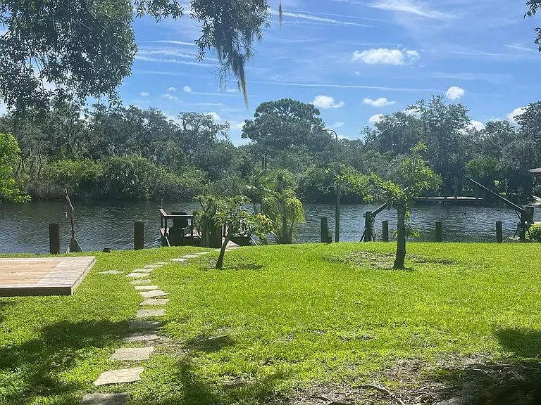 Waterfront backyard with open lawn, mature shade trees, and a path leading to a private dock on the water.