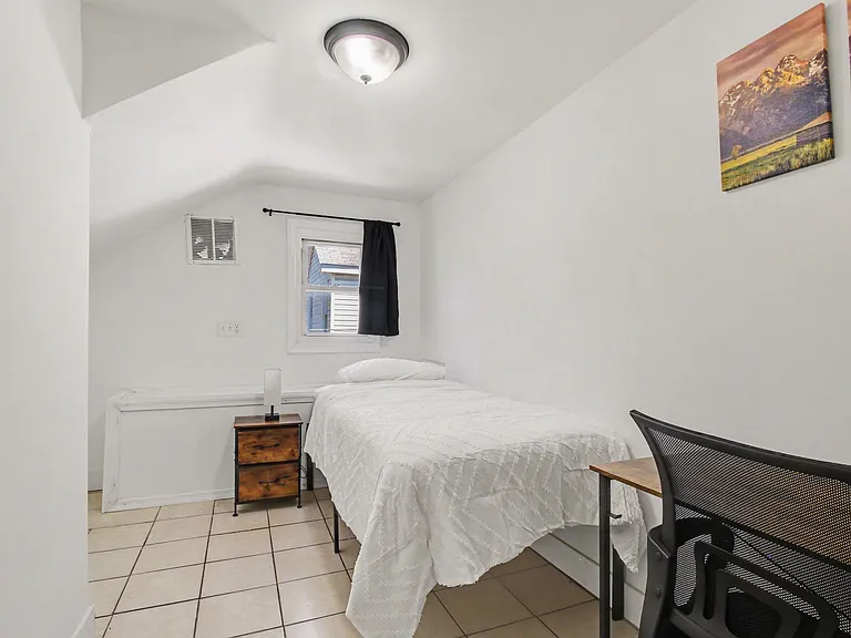 Private bedroom with a furnished bed, nightstand, and desk setup, plus a window for natural light.