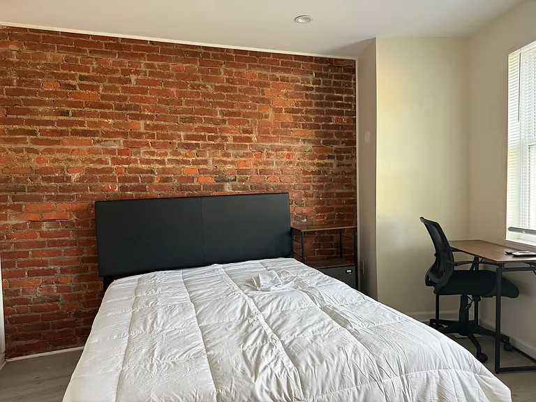 Furnished bedroom with exposed brick accent wall, comfortable bed, and dedicated desk space beside a window with blinds.