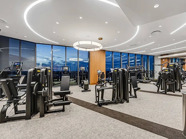 24-hour fitness center with weight machines, treadmills, ellipticals, stationary bikes, a squat rack, a punching bag, medicine balls, and two full walls of windows overlooking the city.