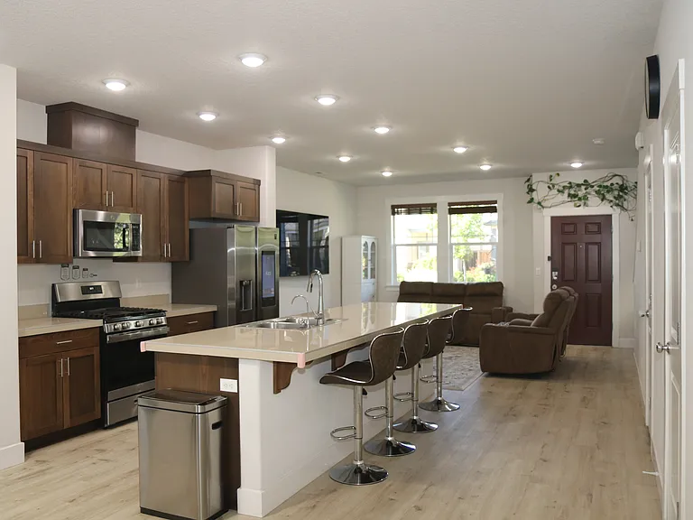 Open_concept kitchen and living space featuring stainless steel appliances, 12 feet island with seating, vinyl flooring, and bright recessed lighting. Living area with large windows for natural light, and a welcoming front entry.