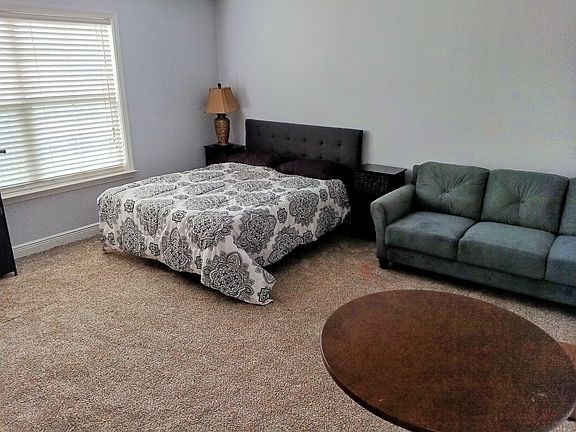 Spacious furnished bedroom with a full bed, nightstands, and an in-room sofa, plus a large window with blinds.