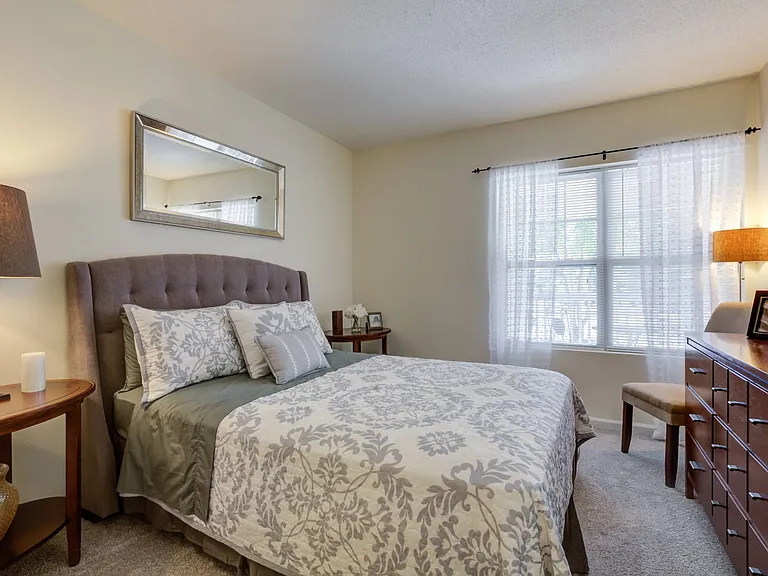 Furnished bedroom with plush carpeting and oversized window with mini-blinds. Room includes queen-sized bed, two nightstands, chair, lamp and horizontal dresser. at Summit Place apartments in Methuen,