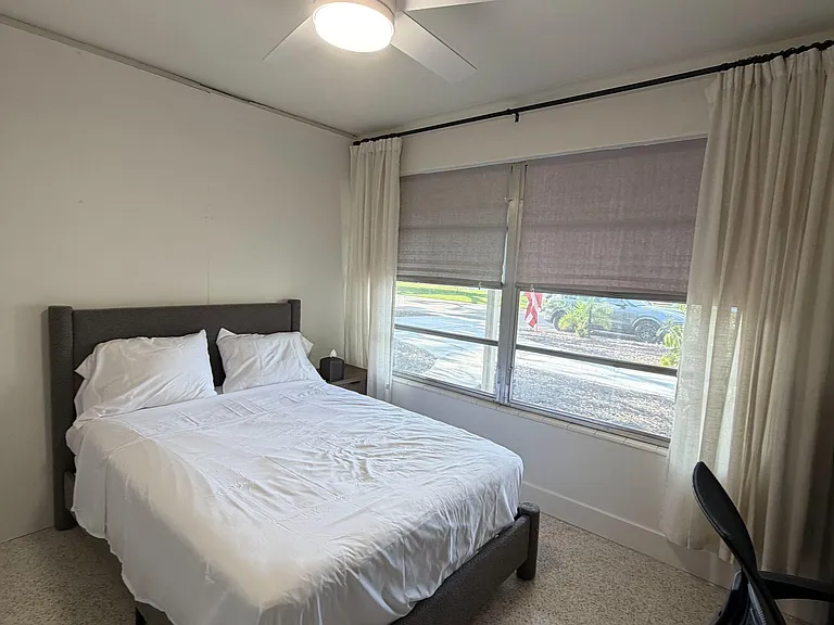 Furnished bedroom with a full bed, ceiling fan, and large window with curtains and shades for light control.