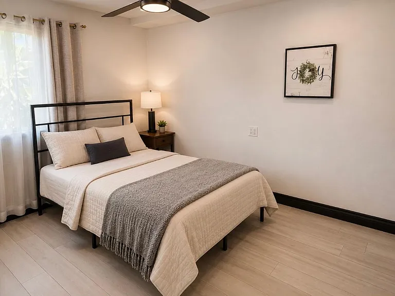 Bedroom features a queen bed, nightstand with lamp, ceiling fan, and a window with layered curtains for privacy.