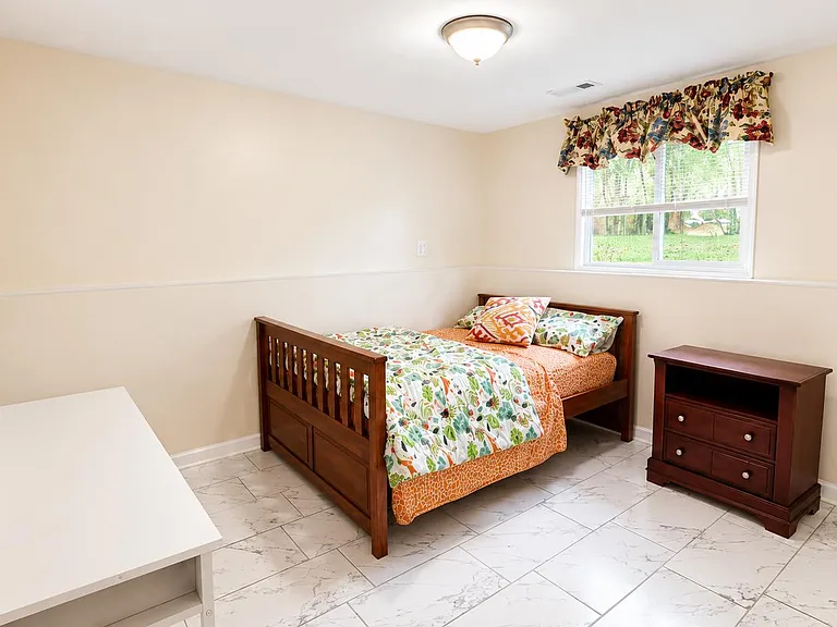 Furnished bedroom with a twin bed, dresser, and desk area, plus a large window overlooking the yard.