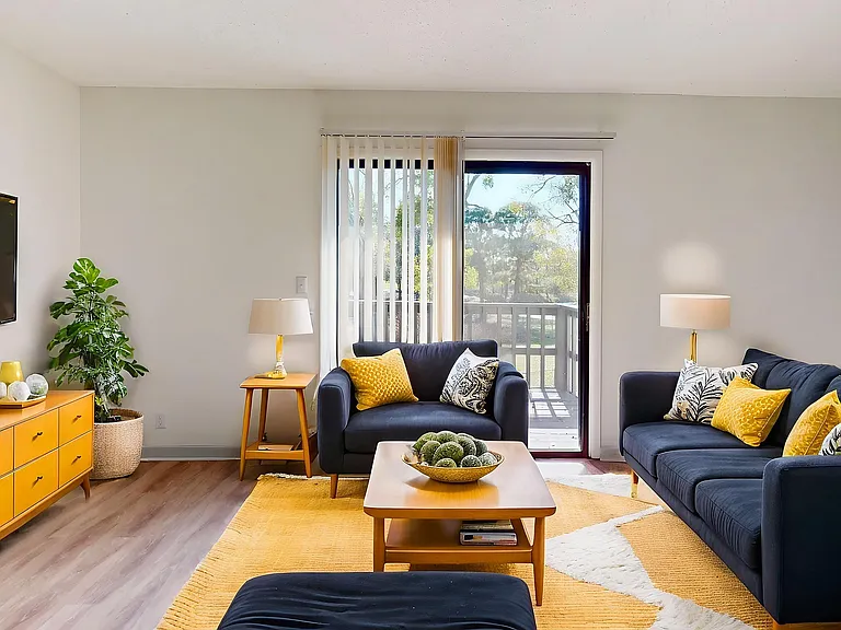 Modern living room with stylish navy sofas, vibrant yellow accents, and abundant natural light, enhanced by a large sliding glass door leading to a scenic view. Elegant decor includes a sleek TV, contemporary lamps, and a lush indoor plant.