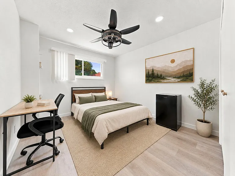 Furnished bedroom with a work desk, ceiling fan, window, and a personal mini fridge for added convenience.